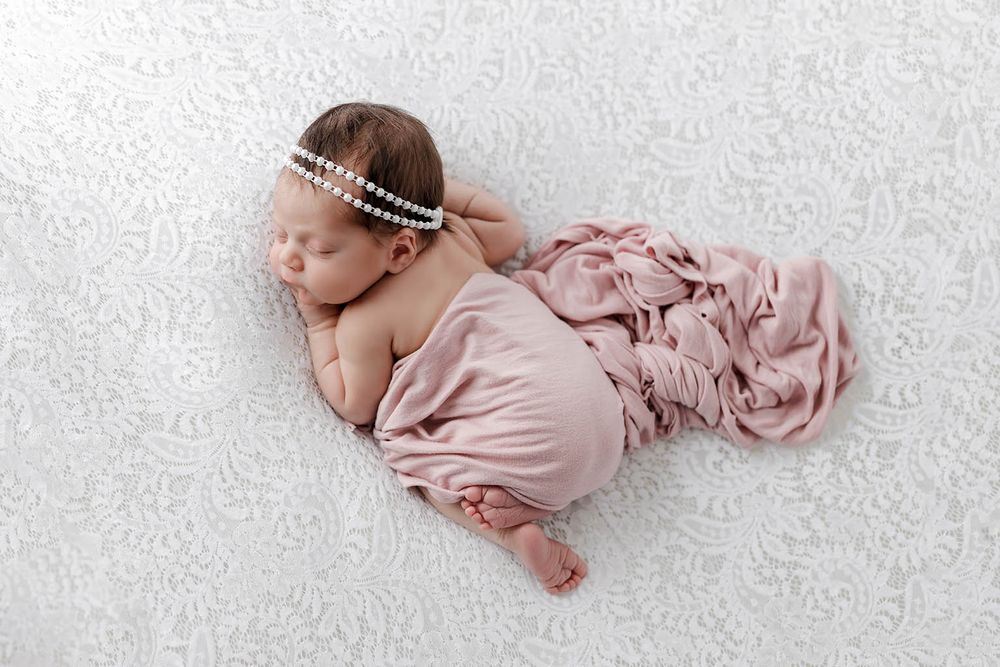 Newborn baby girl posed in pink wrap with pearl headband on white backdrop.
