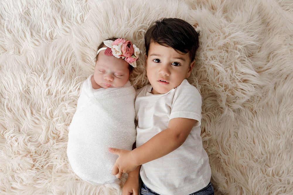 Newborn baby girl in white swaddle with floral headband posed with toddler brother.