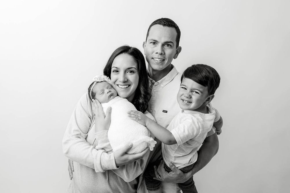 Black and white family portrait of smiling parents holding newborn baby and toddler against white background.