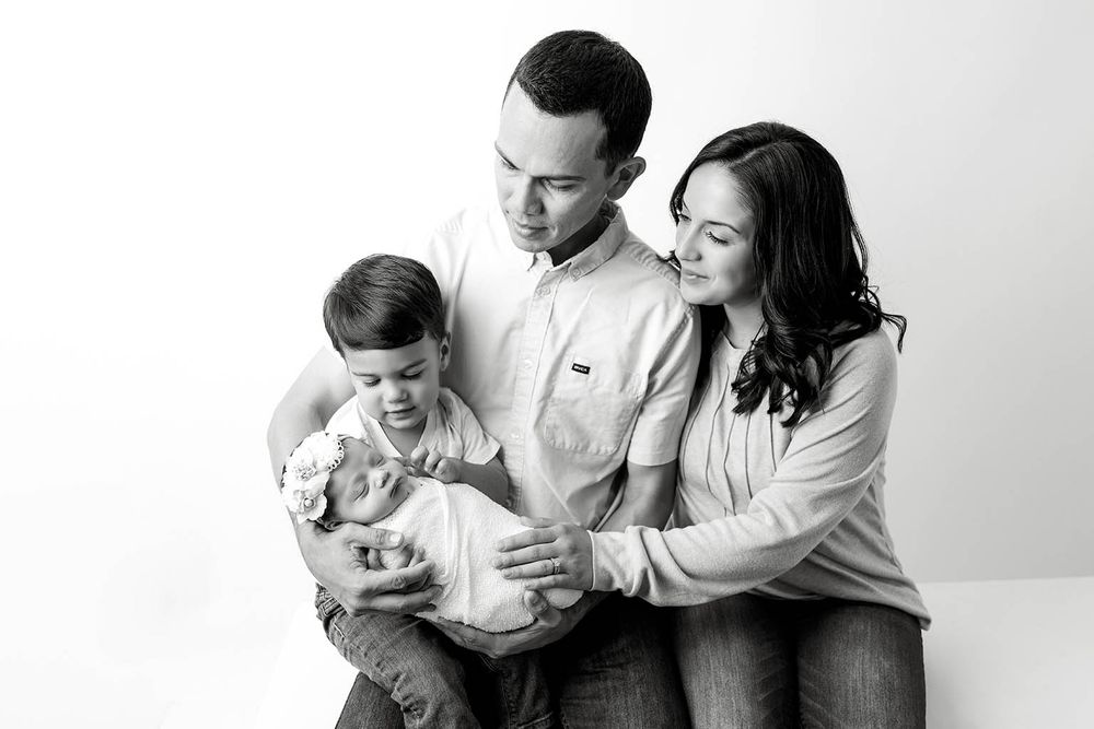Black and white family portrait with parents and toddler admiring newborn baby in studio setting.
