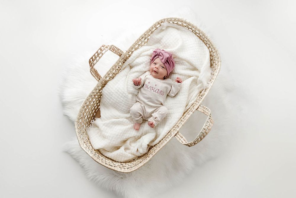 Newborn baby in personalized white outfit with pink bow headband sleeping in a wicker Moses basket.