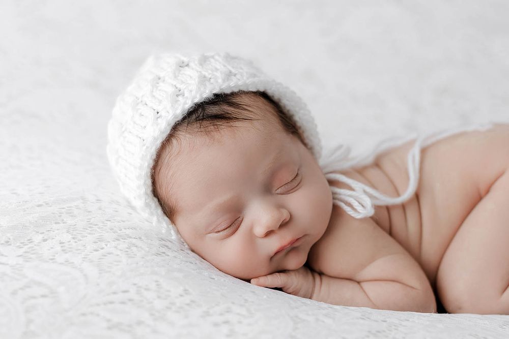 A peaceful sleeping newborn baby wearing a white knit bonnet rests on a soft white lace blanket.