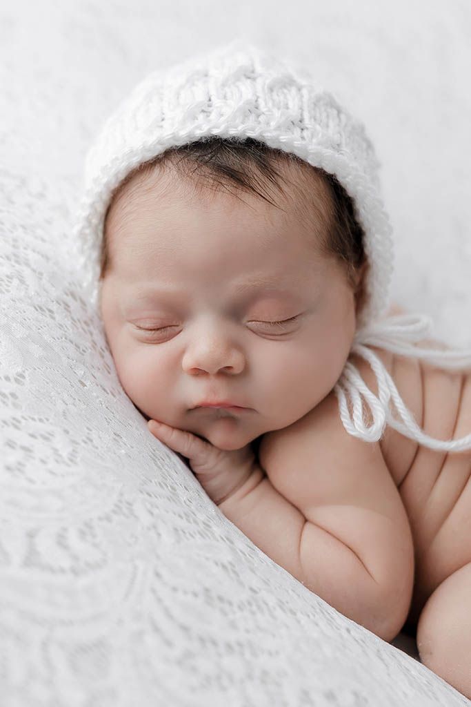 Close-up portrait of a sleeping newborn baby in a white knit bonnet resting on a white lace blanket.