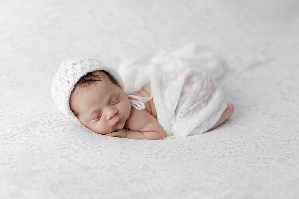 Peaceful sleeping newborn baby wrapped in white lace swaddle wearing knit bonnet on white backdrop.