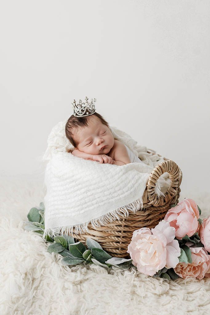 Sleeping newborn baby wearing a tiny crown resting in a wicker basket with white blanket and pink flowers.