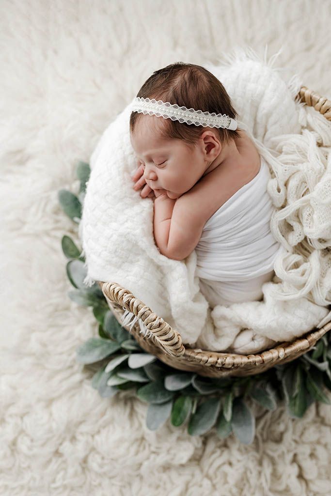 Sleeping newborn baby girl wrapped in white swaddle with lace headband in wicker basket with greenery.