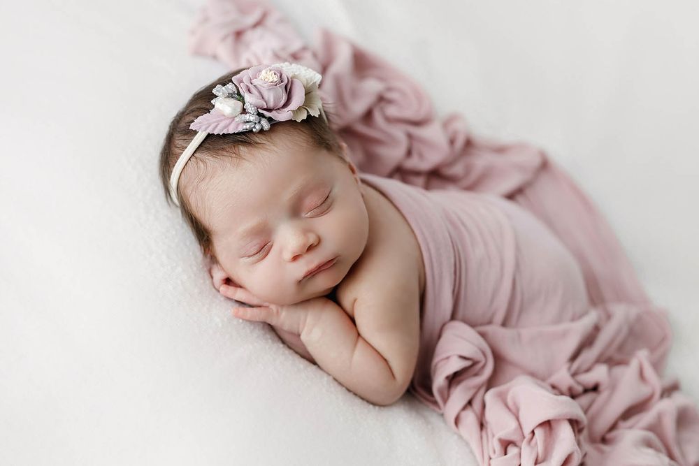 Newborn baby wrapped in dusty pink with a floral headband sleeping on white fur.