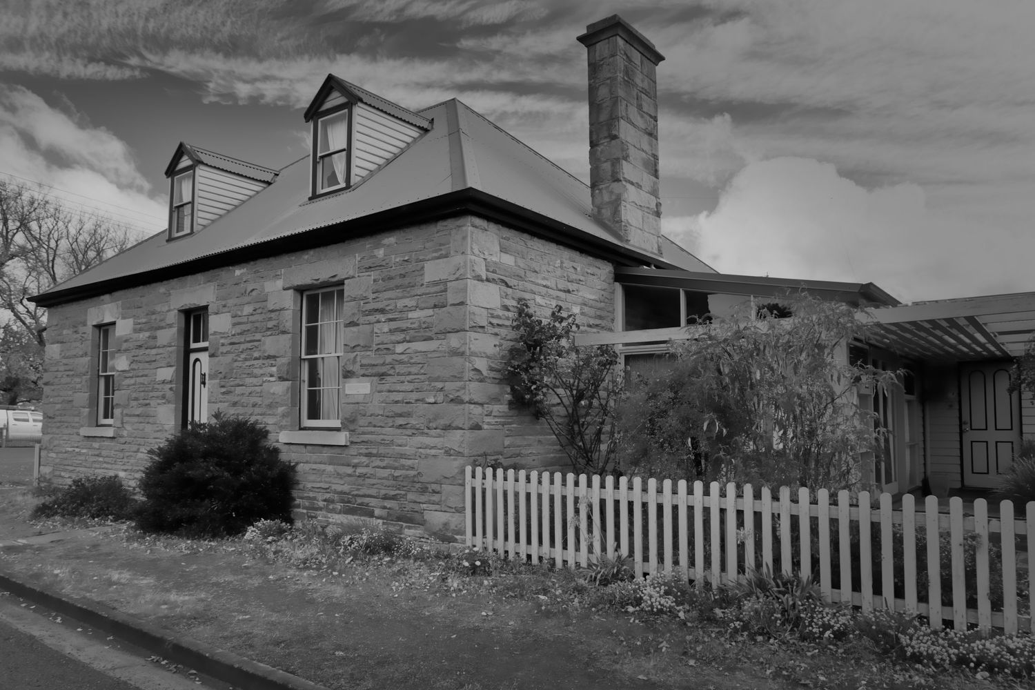 Black and white photo of historic stone cottage with white picket fence and chimney.