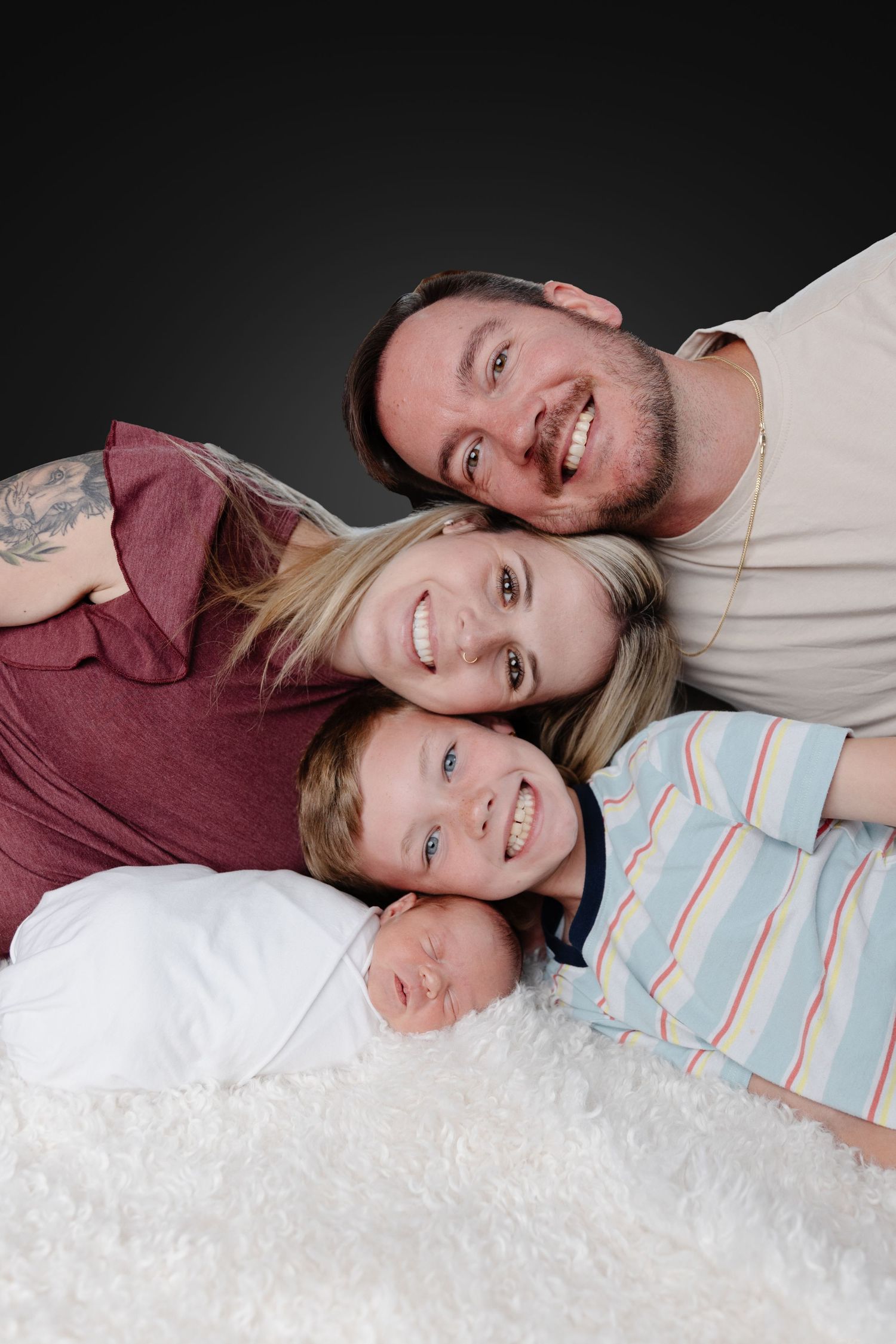 A smiling family of four poses together on a white fluffy rug, including parents, a young boy, and a newborn baby.