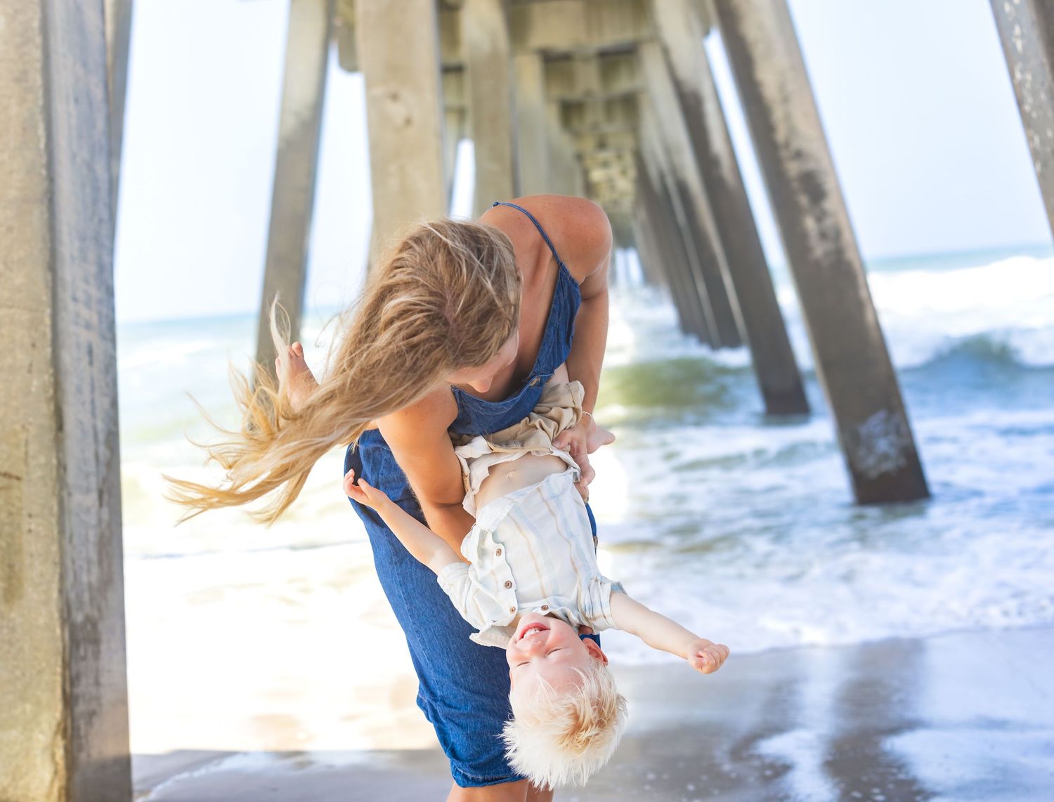 A mother joyfully lifts her toddler on a sunny beach beneath a wooden pier with ocean waves in the background.