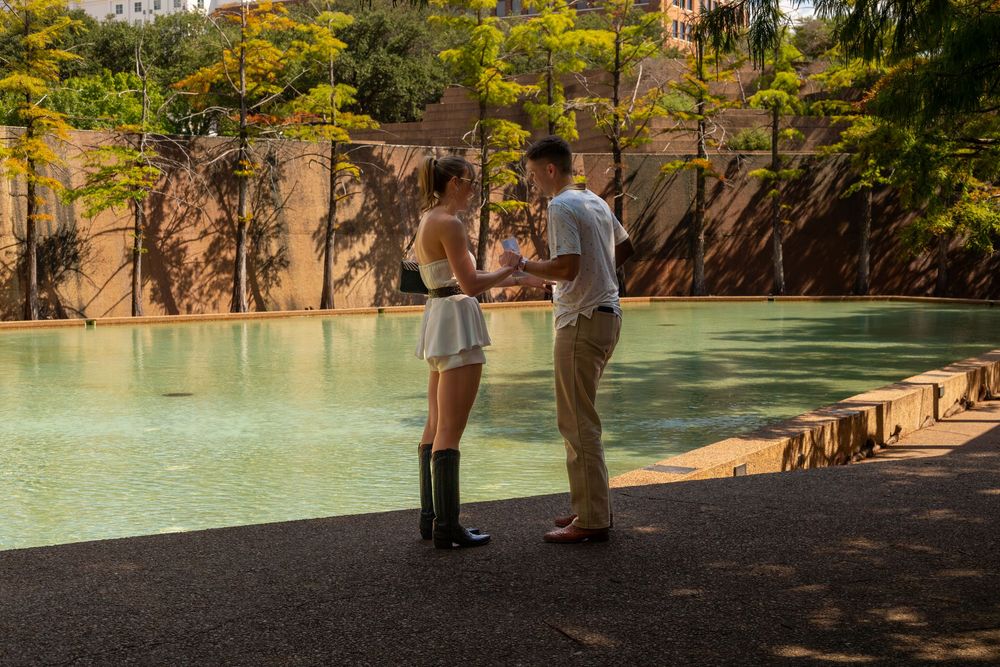 Couple standing by a serene green pool surrounded by lush trees and rocky walls in an outdoor setting.