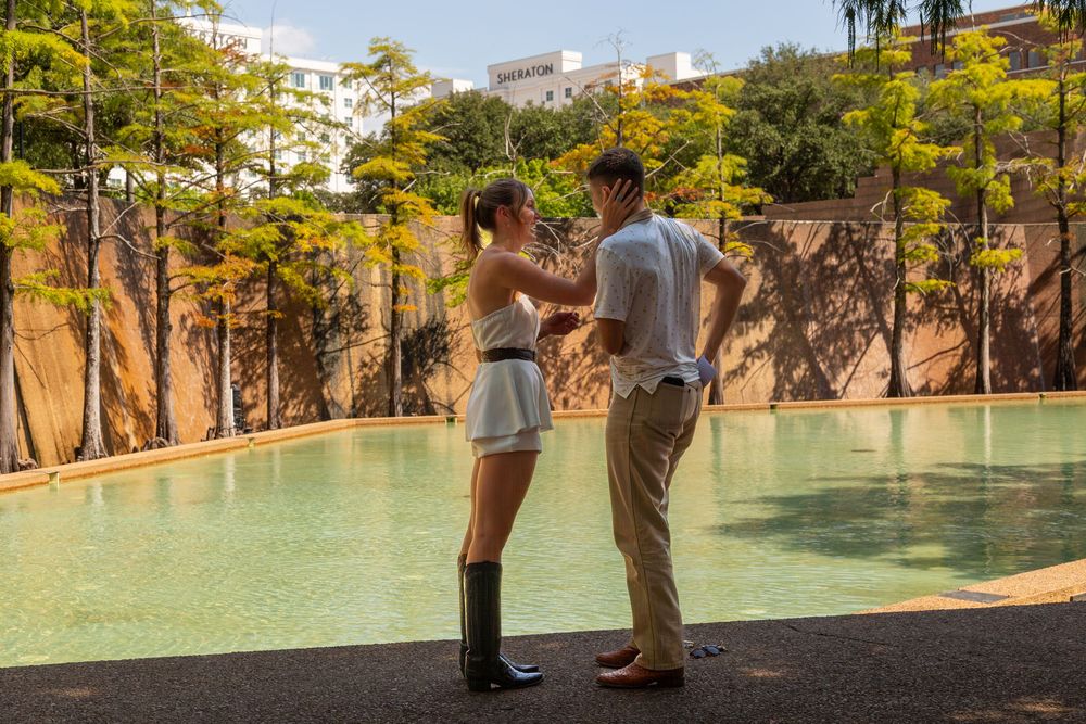 Couple dancing outdoors near a serene pool surrounded by lush green trees in a tropical urban setting.