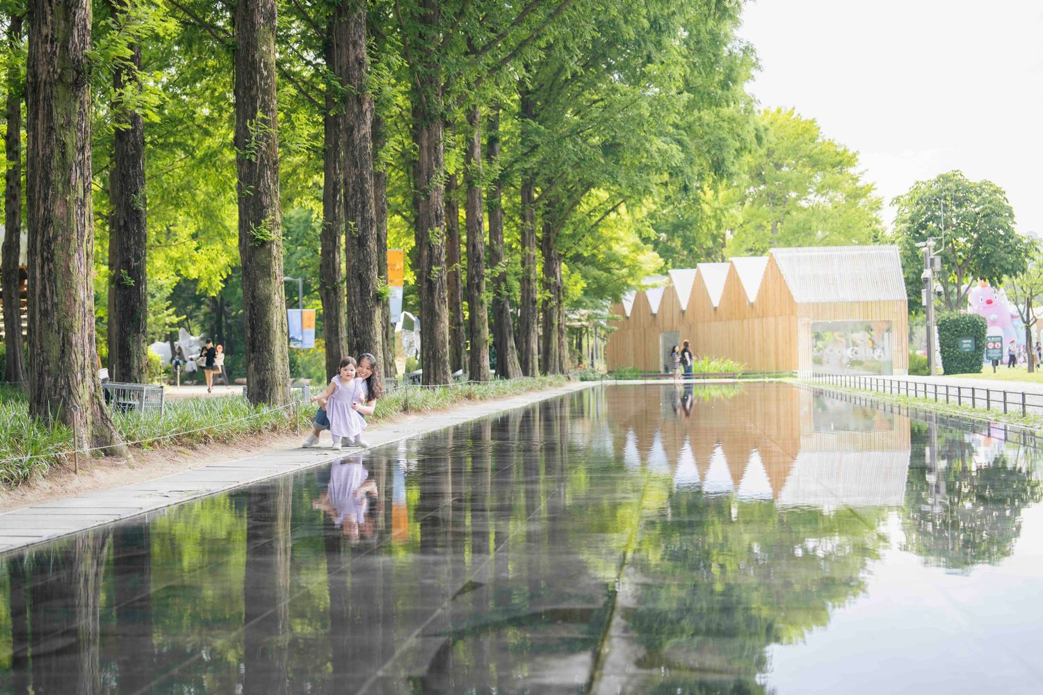 Serene park reflecting pool mirrors tall green trees and wooden pavilion structures on a sunny day.