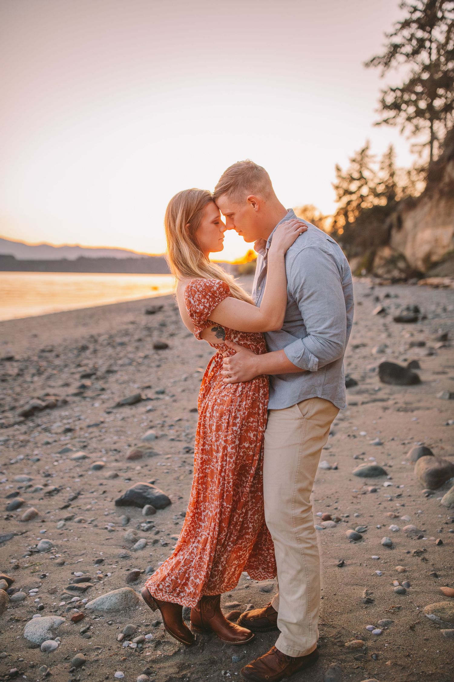 Couple embracing on rocky beach at golden sunset, woman in red floral dress and man in light blue shirt.