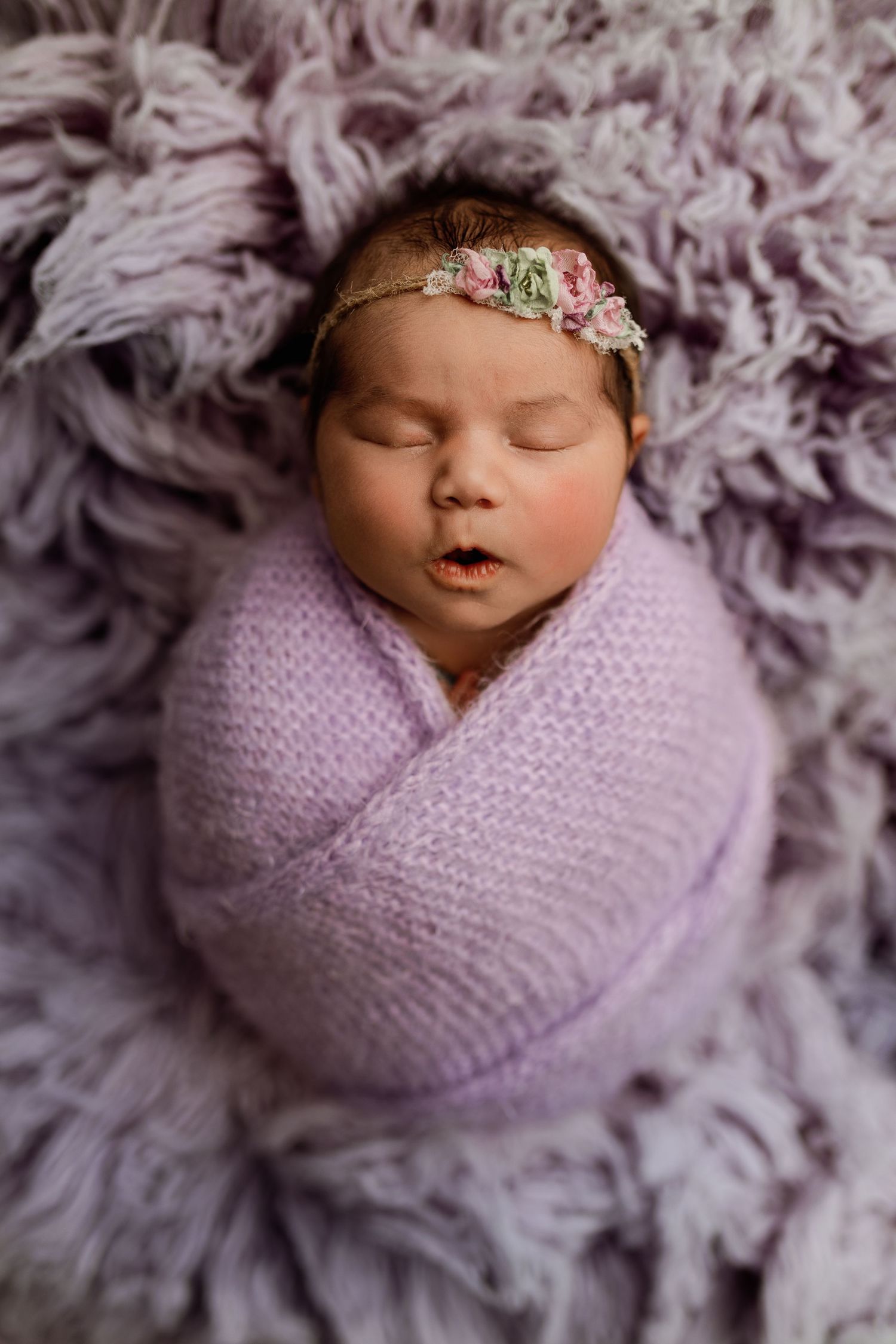 Newborn baby girl swaddled in lavender knit wrap with floral headband, sleeping peacefully on purple fluffy blanket.
