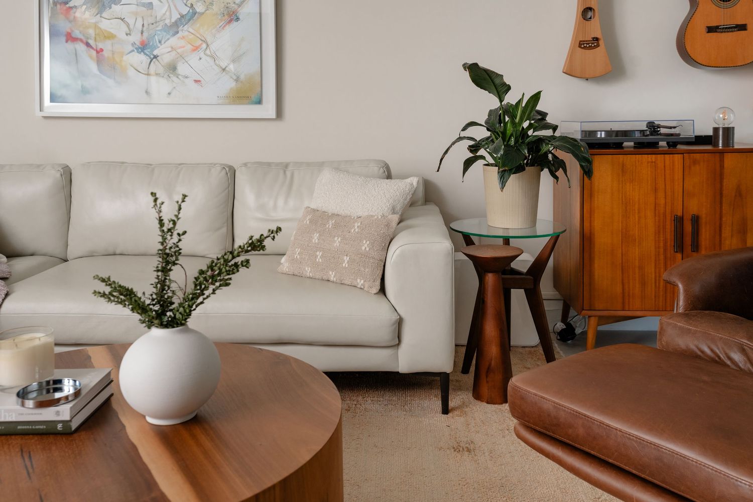 Modern living room with white leather sofa, wooden coffee tables, indoor plants, and a vintage wooden cabinet with guitars.