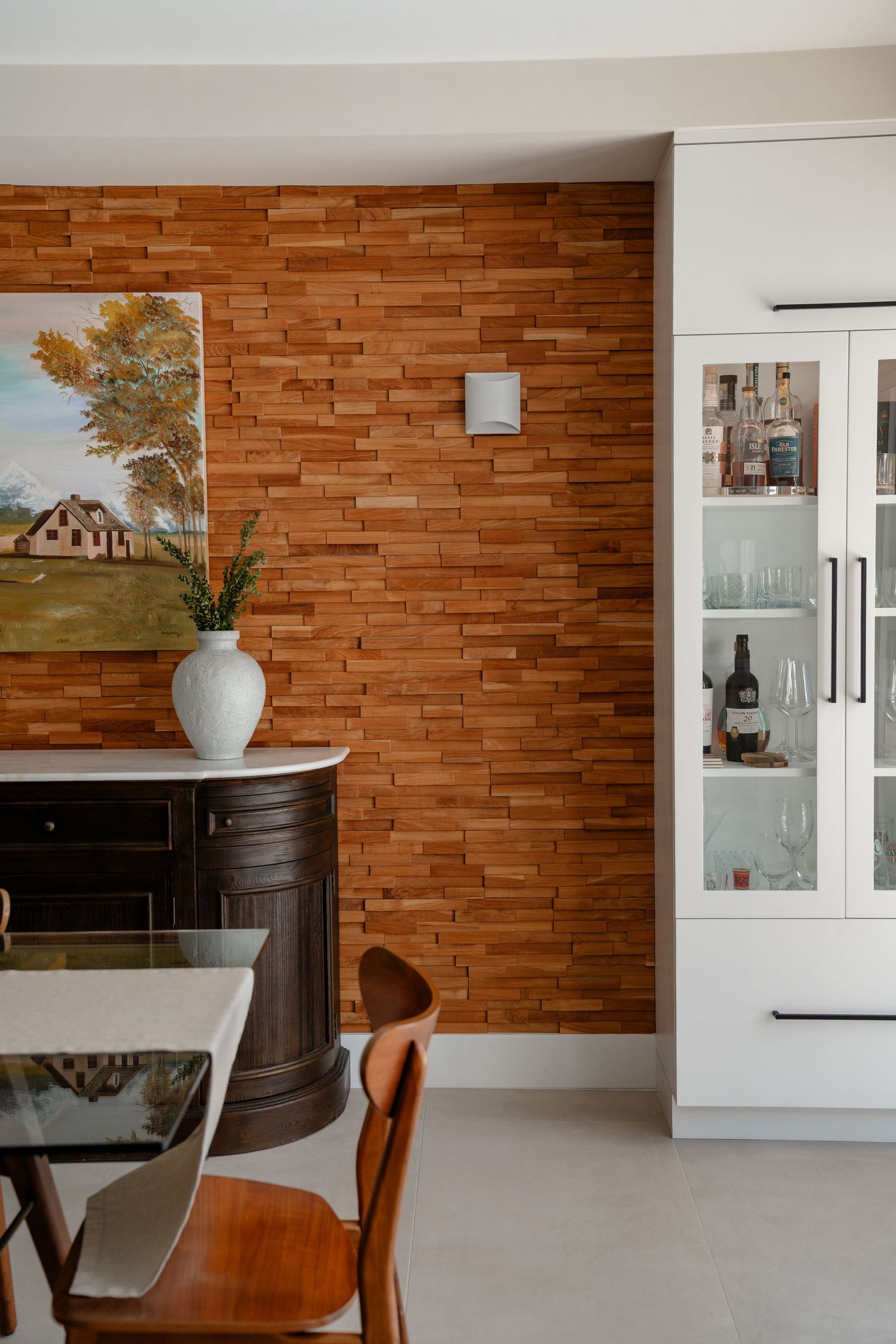 Dining area with wood panel accent wall, dark sideboard, white vase, glass cabinet with bottles, and wooden chairs.
