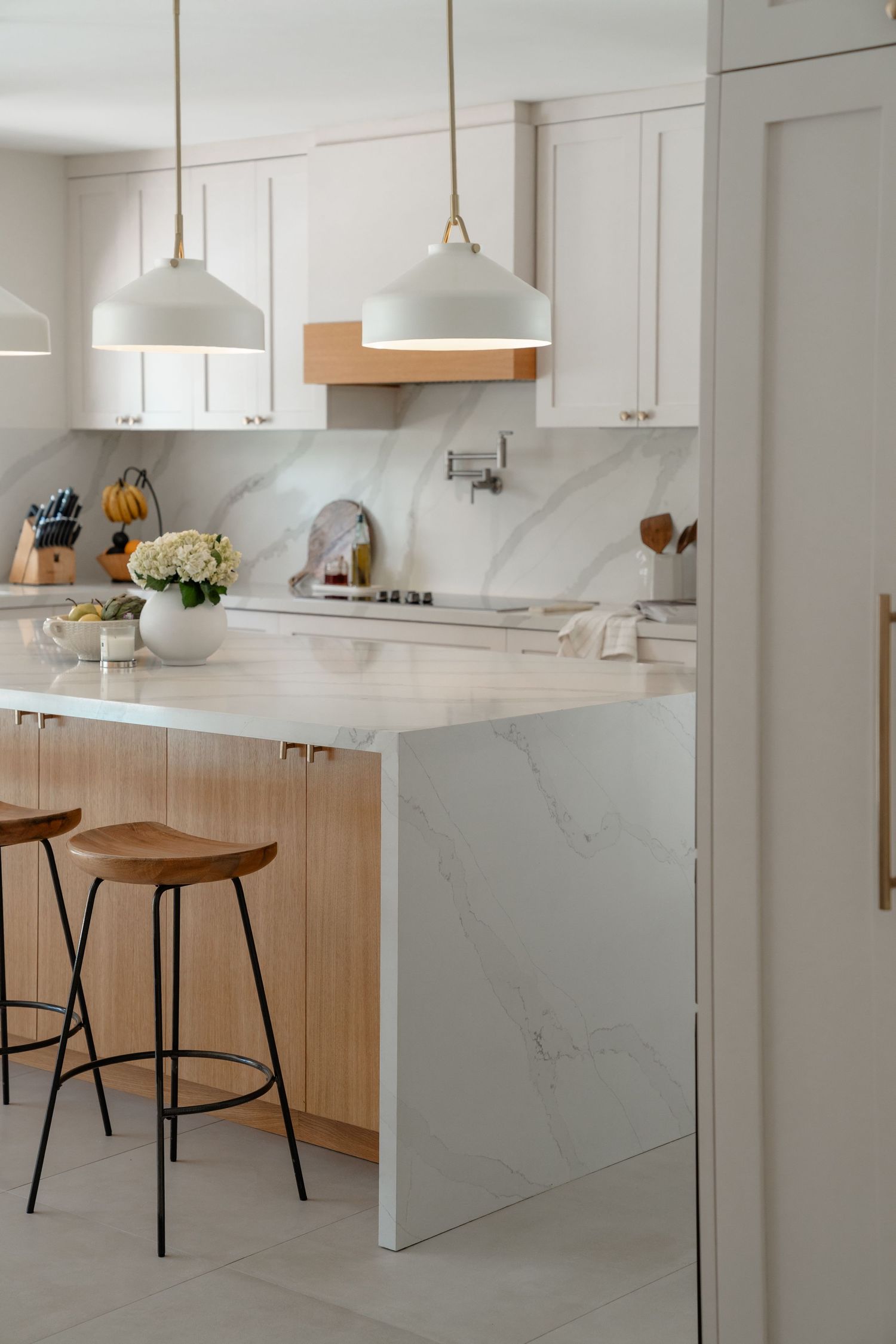 Modern white kitchen with marble island, wood bar stools, pendant lights, and shaker cabinets.