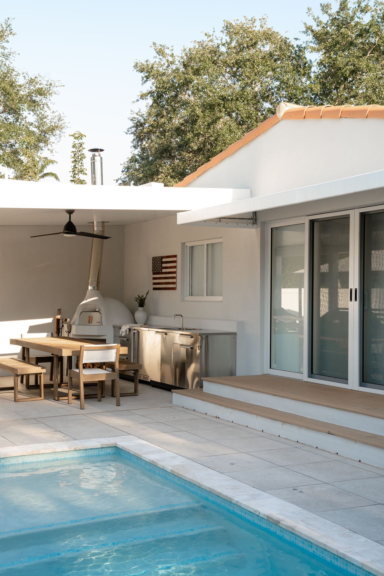 Modern outdoor kitchen with pool, wood dining table, pizza oven, and stainless steel grill beside a white stucco home.