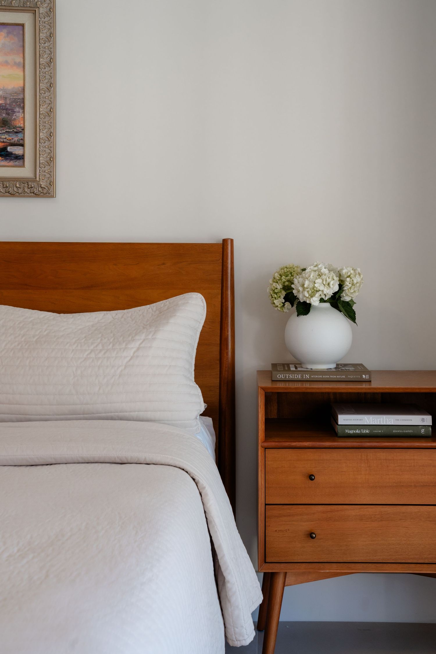 Mid-century modern bedroom with warm wood bed frame, white bedding, matching nightstand, books, and white hydrangea arrangement.