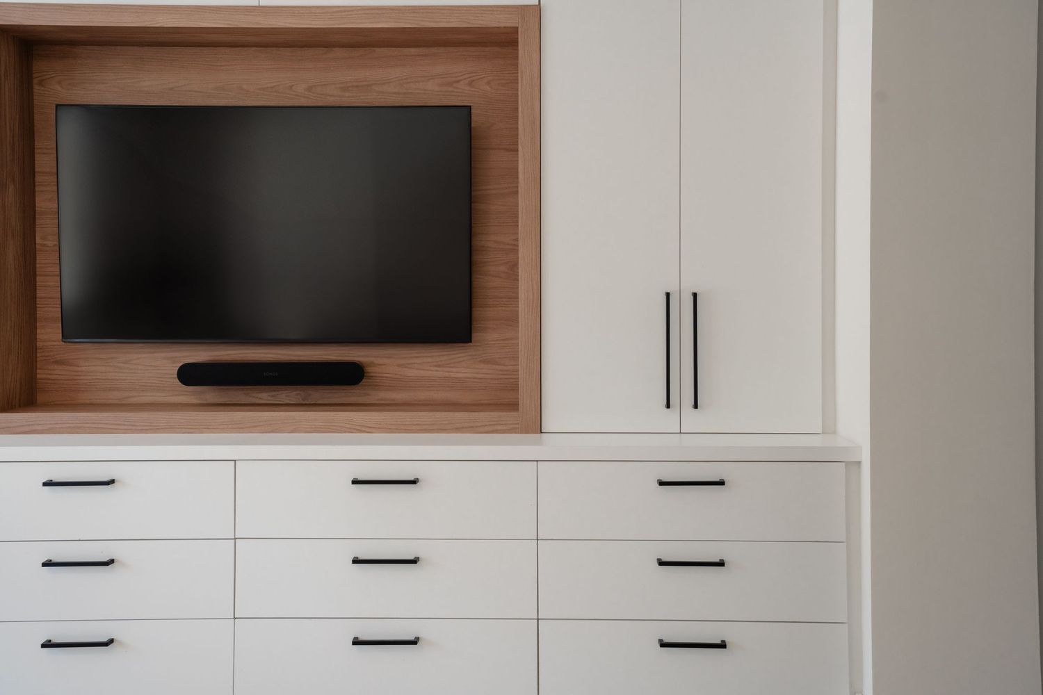 Modern built-in white cabinetry with black handles, featuring a wooden TV niche with a flat-screen TV and soundbar.