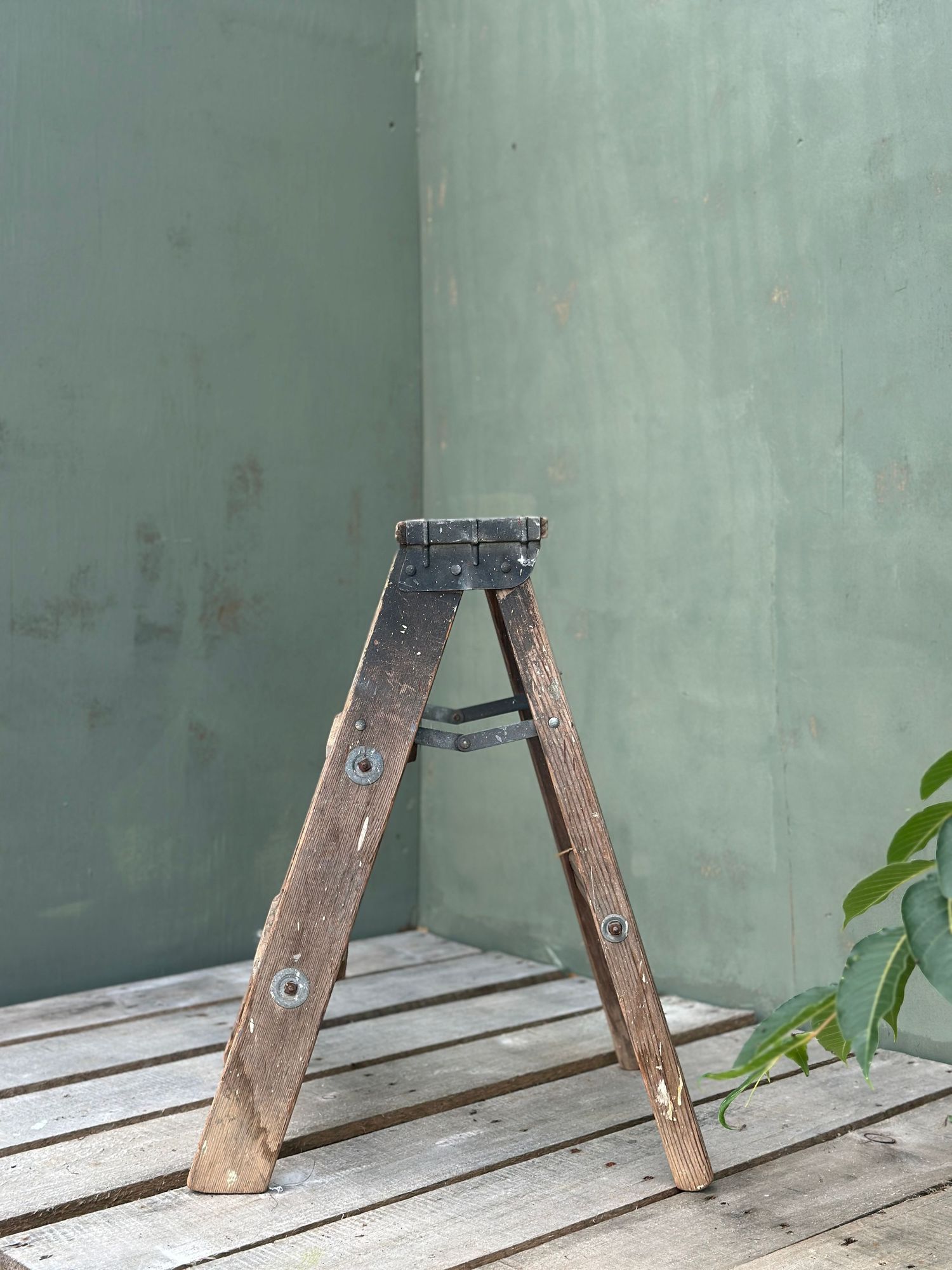 Vintage wooden A-frame step ladder leaning against a green wall on rustic wooden deck flooring.