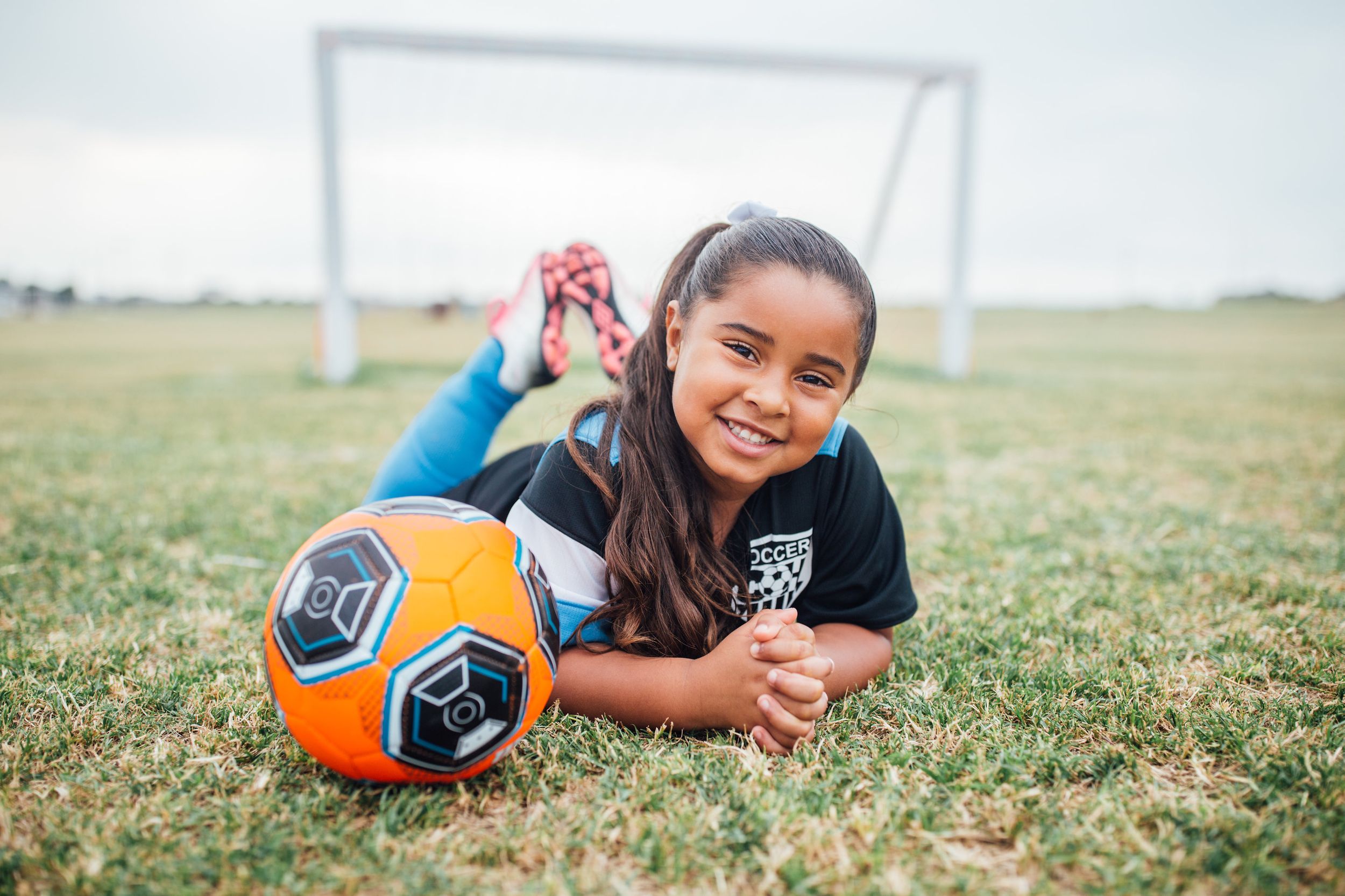 portrait of small girl with soccer ball laying in grass on stomach, feet up and hands folded, smiling, with soccer net