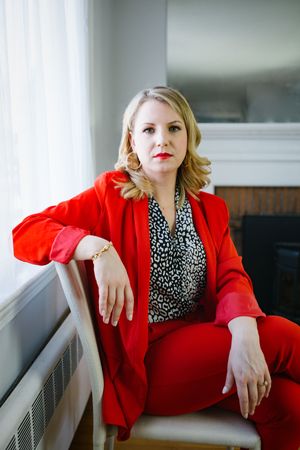 sitting portrait of white woman, blonde curls wearing a red suit pant suit with matching red lipstick in bright light