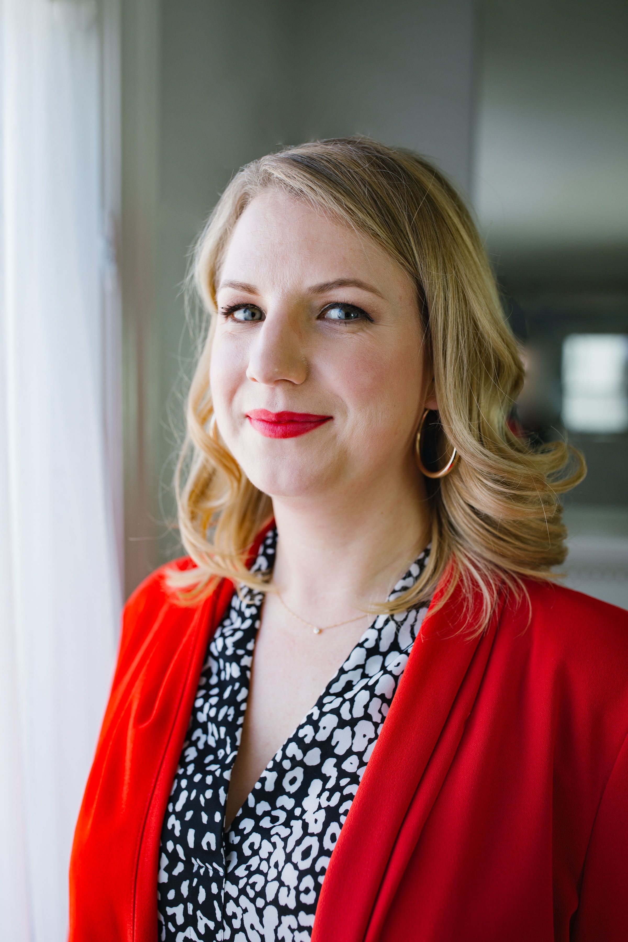 headshot of white woman, blonde curls wearing a red suit pant suit with matching red lipstick, by brightly lit window