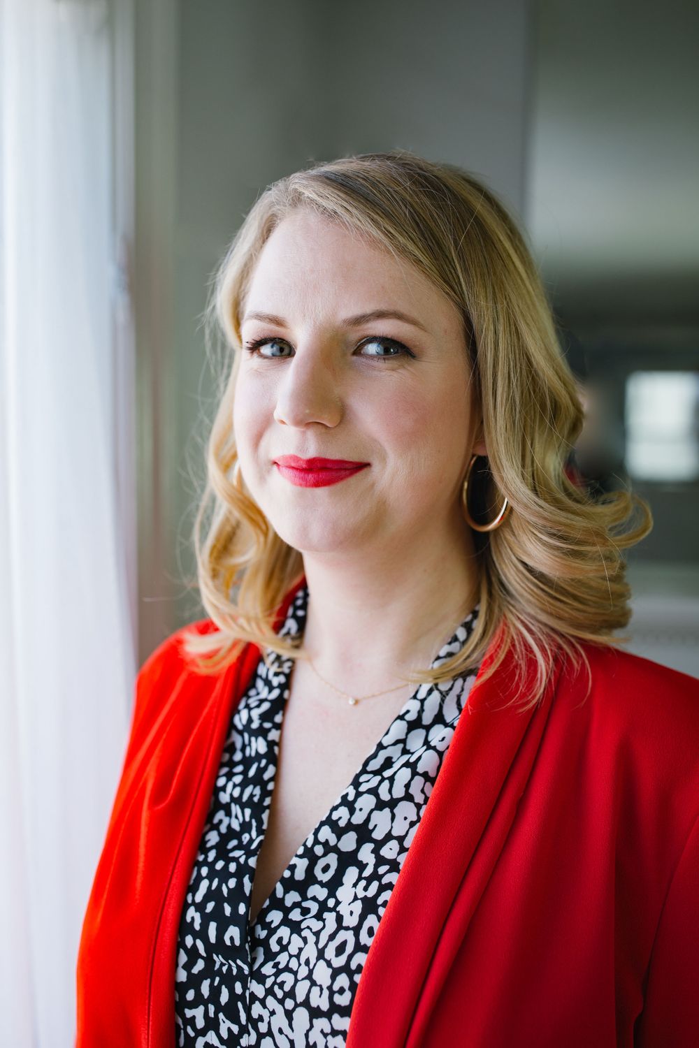 headshot of white woman, blonde curls wearing a red suit pant suit with matching red lipstick, by brightly lit window