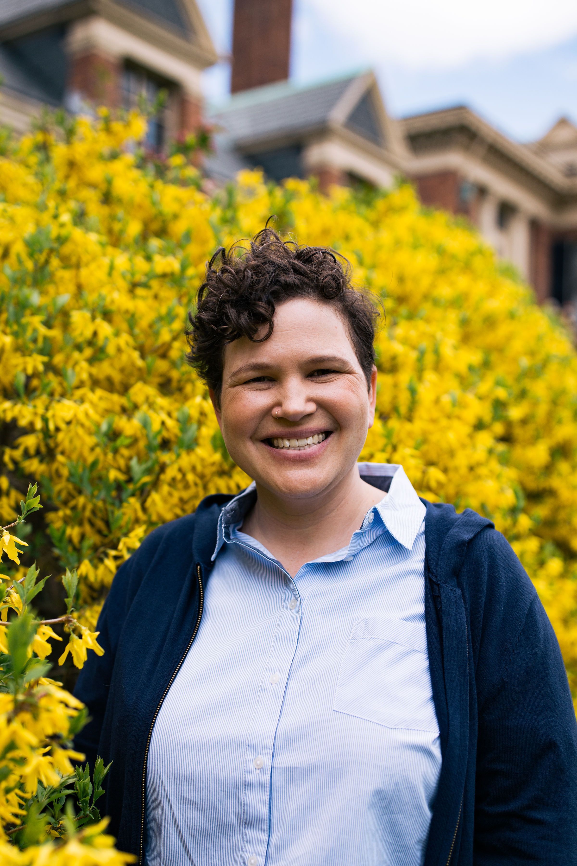headshot image of queer woman smiling, running for local government office, in front of bright yellow Forsythia bush