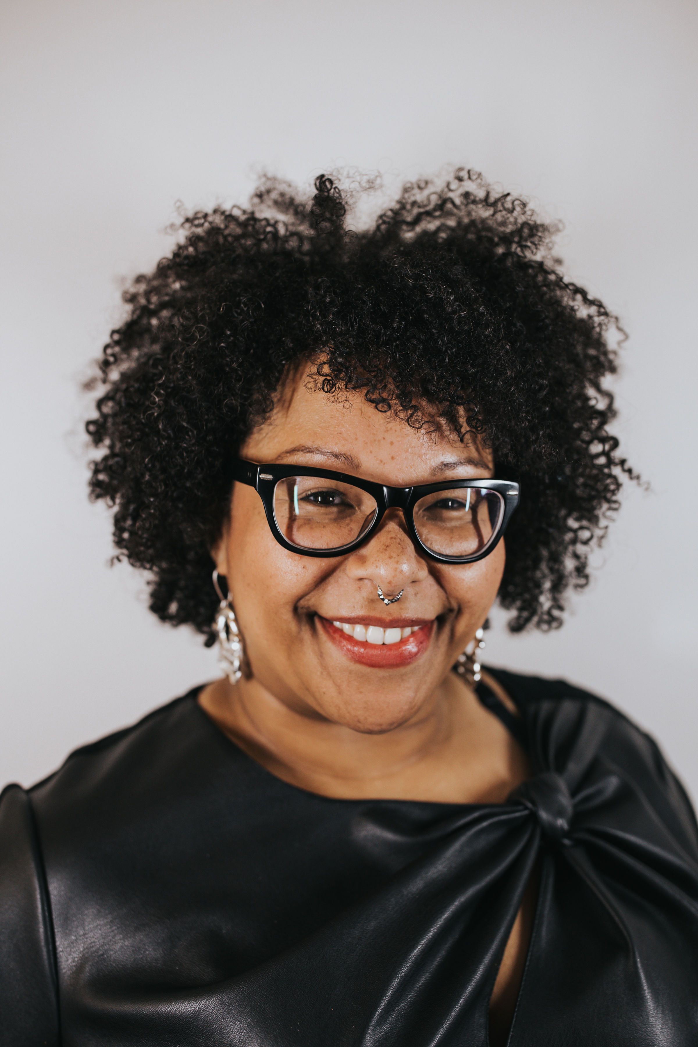 headshot of Black Femme smiling into camera, wearing glasses, septum piercing, natural curly hair, and a red lipstick
