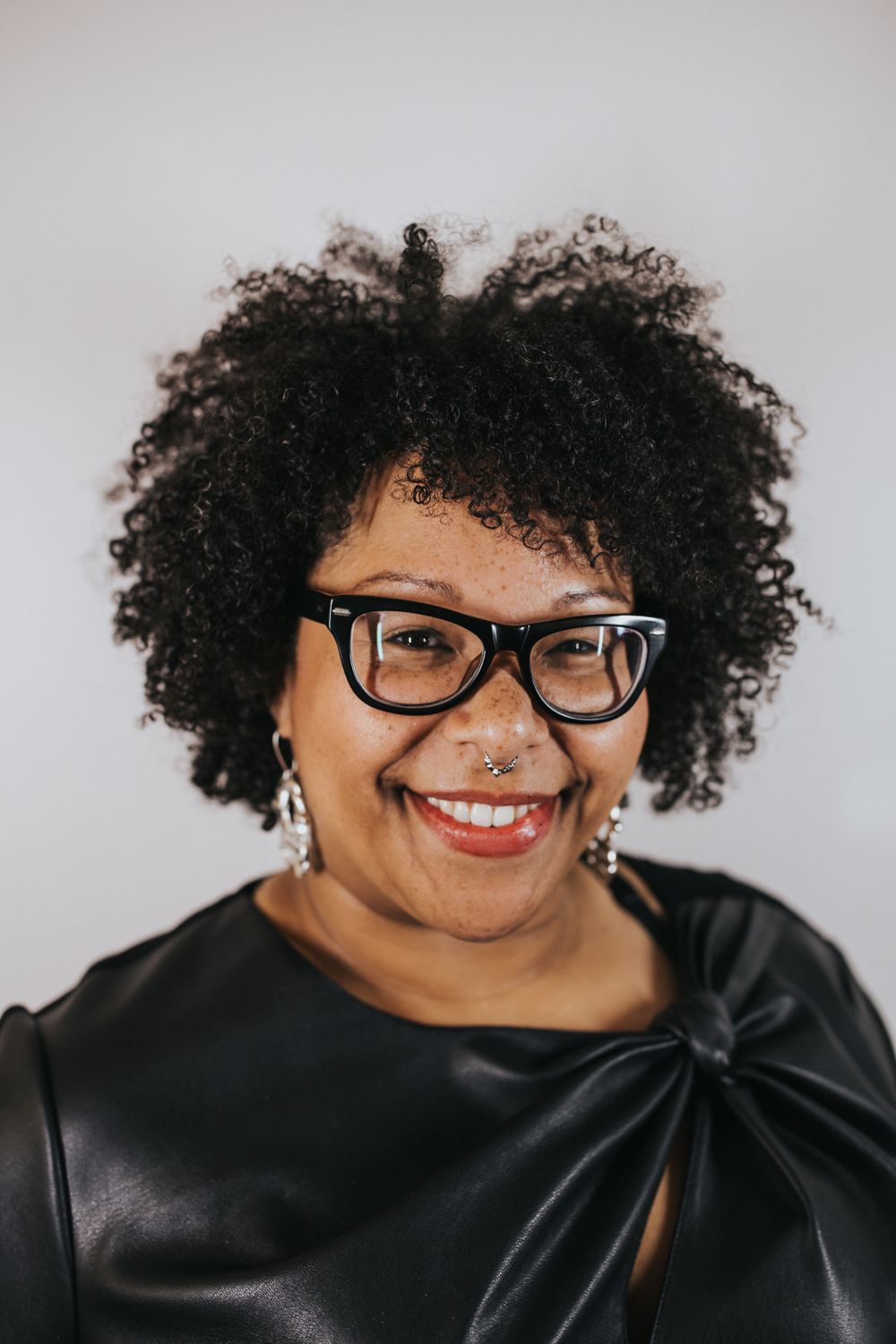 headshot of Black Femme smiling into camera, wearing glasses, septum piercing, natural curly hair, and a red lipstick