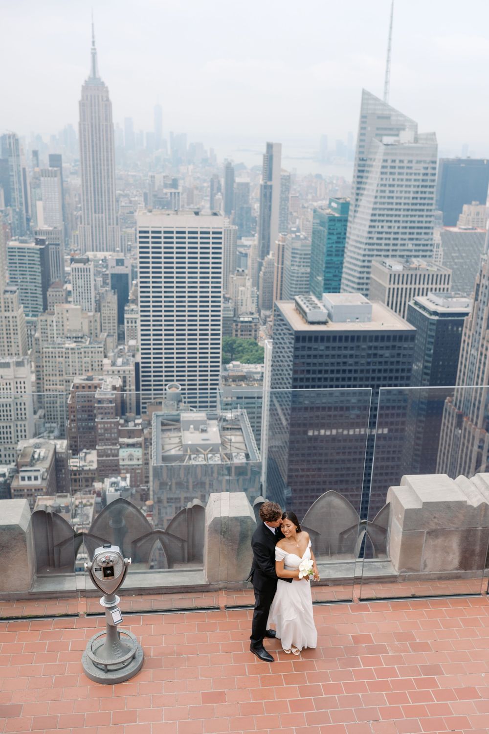 Bride and groom embrace on Top of the Rock observation deck with NYC skyline and Empire State Building backdrop.