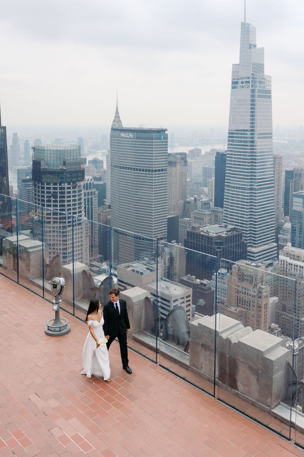 Bride and groom walk on a rooftop observation deck with stunning New York City skyline views on their wedding day.