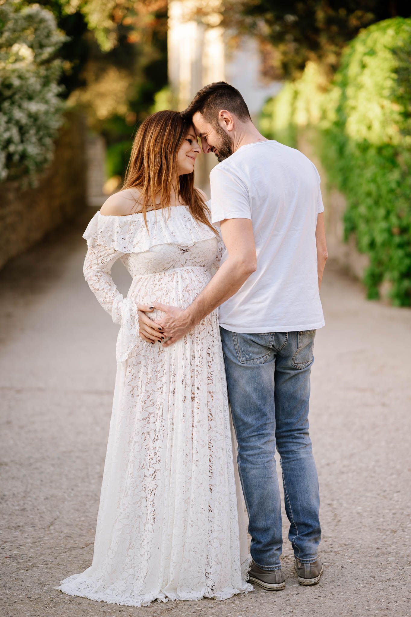 Pregnant couple in white lace maternity gown and jeans embrace tenderly on a sunlit garden path.