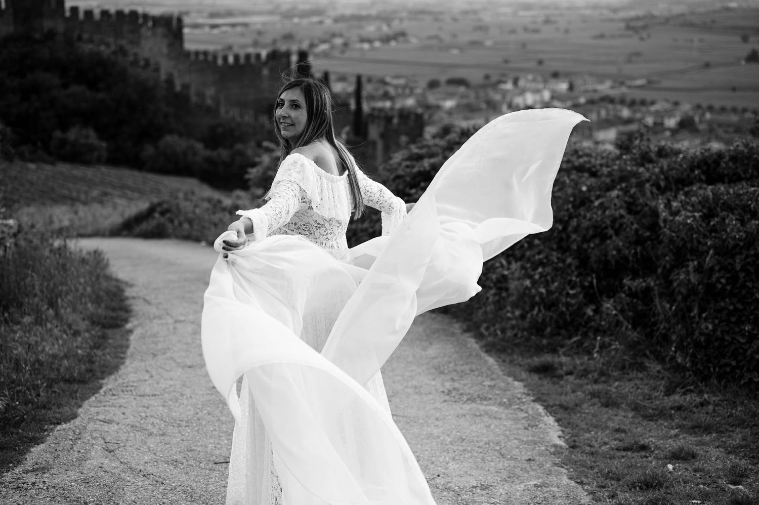Black and white photo of a bride in a flowing white gown, dress billowing in the wind on a scenic path near a castle.