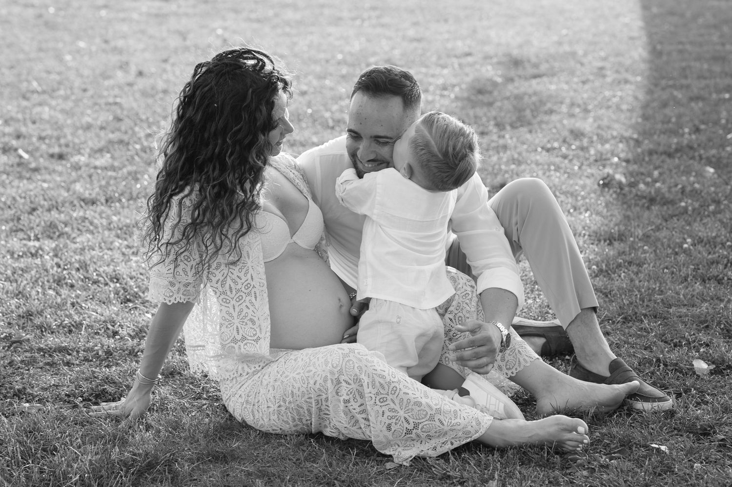 Black and white family photo of parents sitting on grass with toddler, sharing a tender moment outdoors.