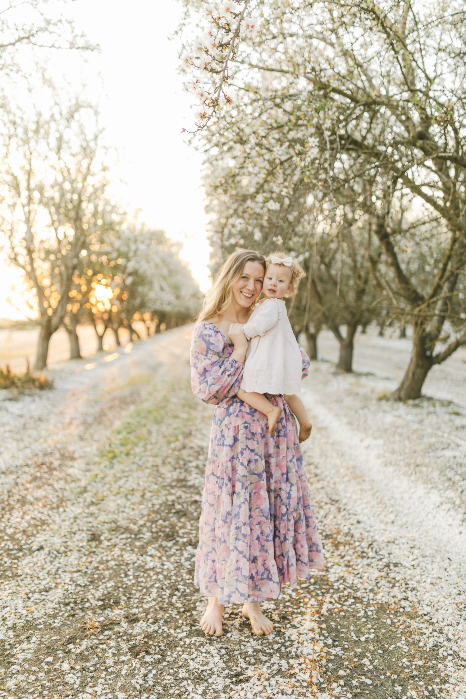 A smiling mother in a floral dress holds her toddler among blossoming trees at golden hour.
