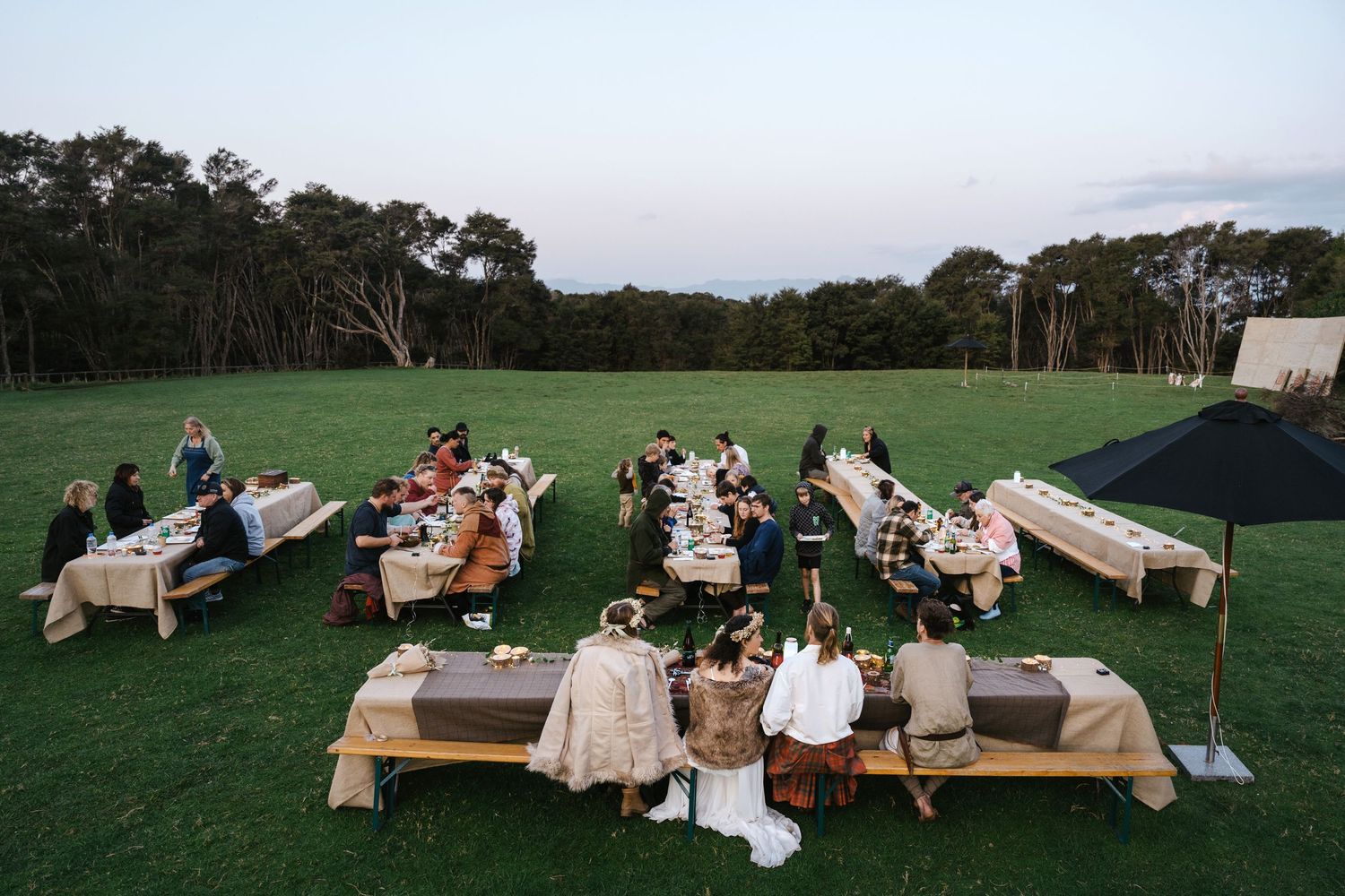 Aerial view of guests dining at long outdoor tables on green grass at dusk, surrounded by trees.