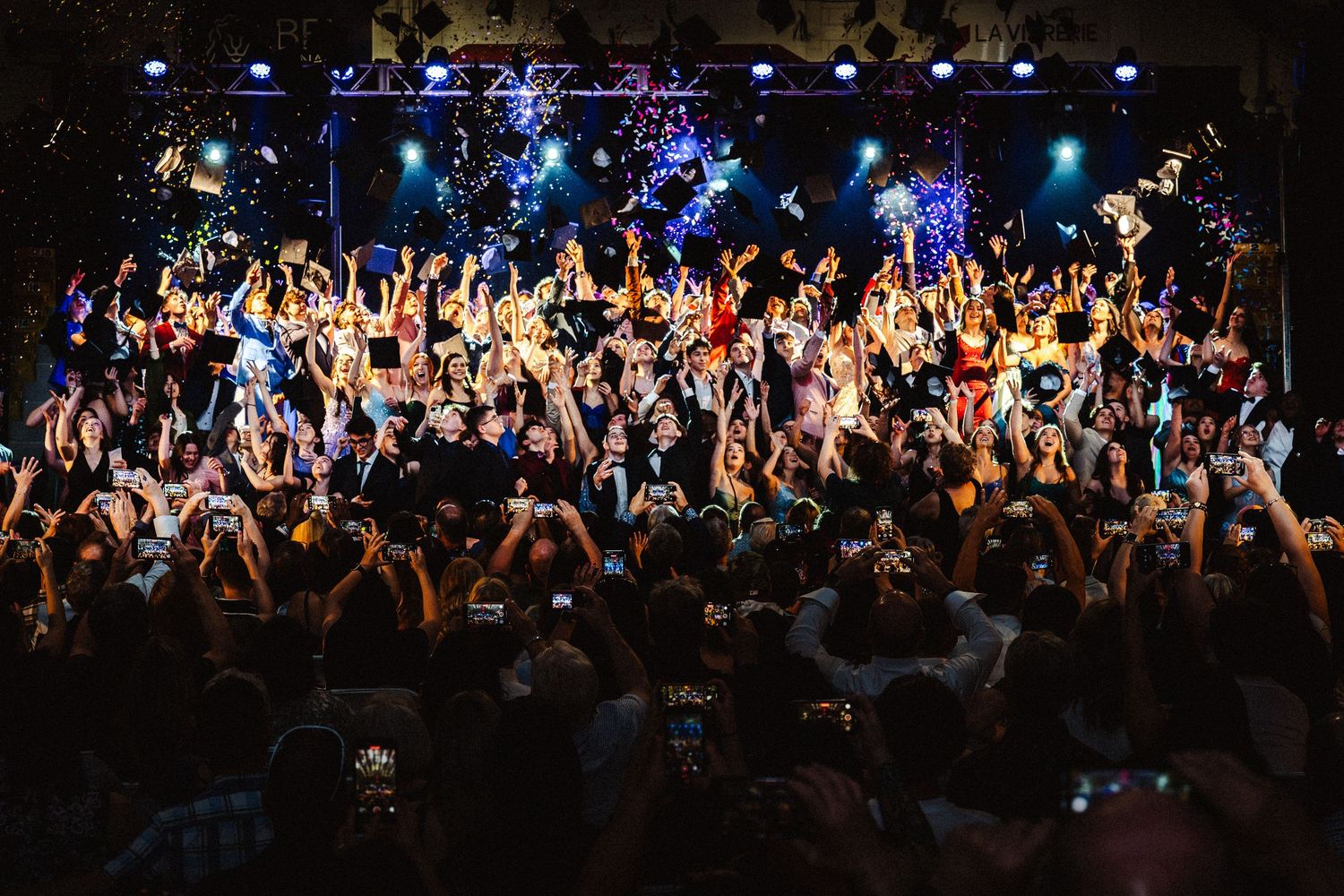 Crowd of concert-goers celebrating with hands raised and confetti falling at a vibrant live music event.