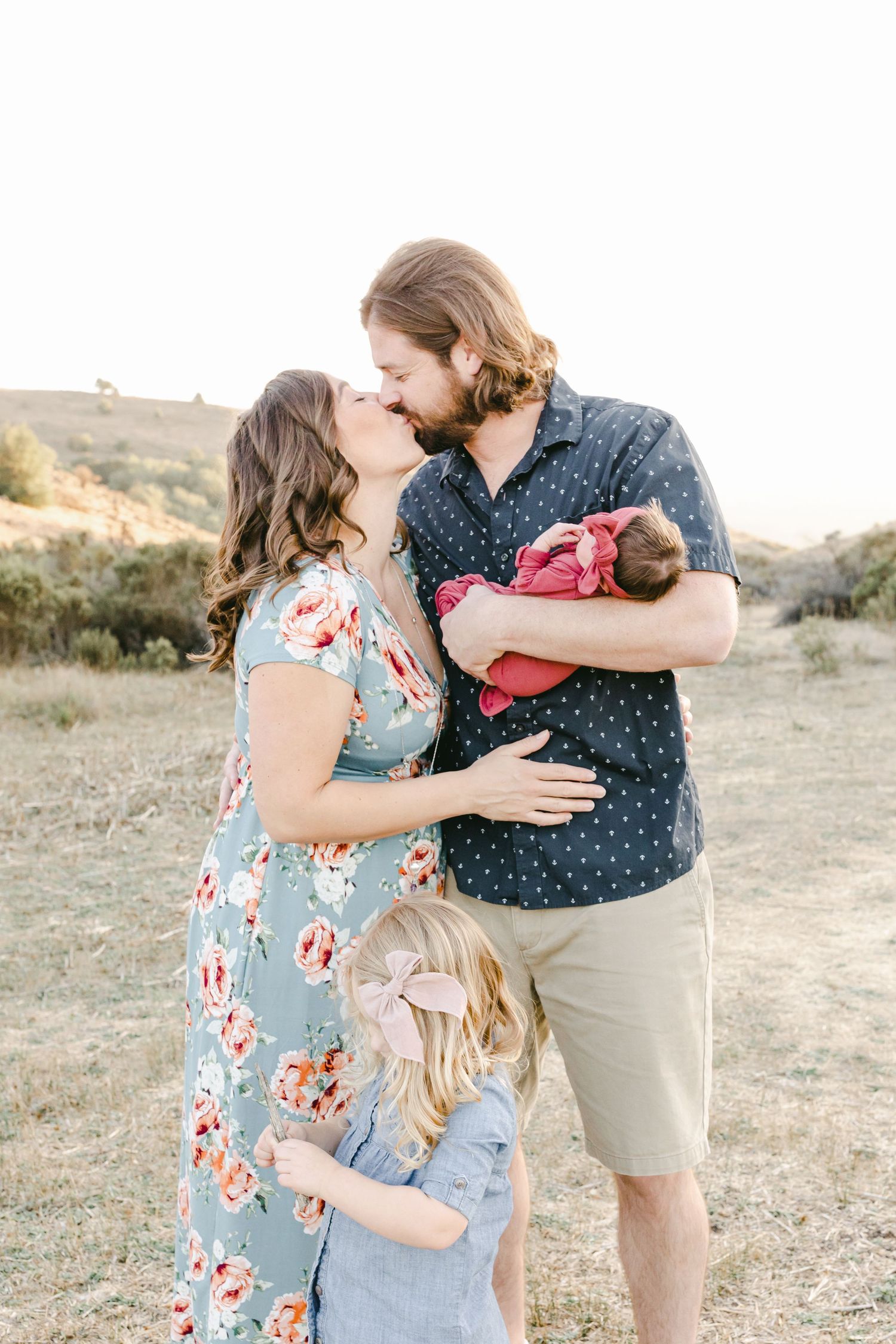 A family of four shares a tender moment outdoors, parents kissing while holding a newborn and a toddler in a sunny field.