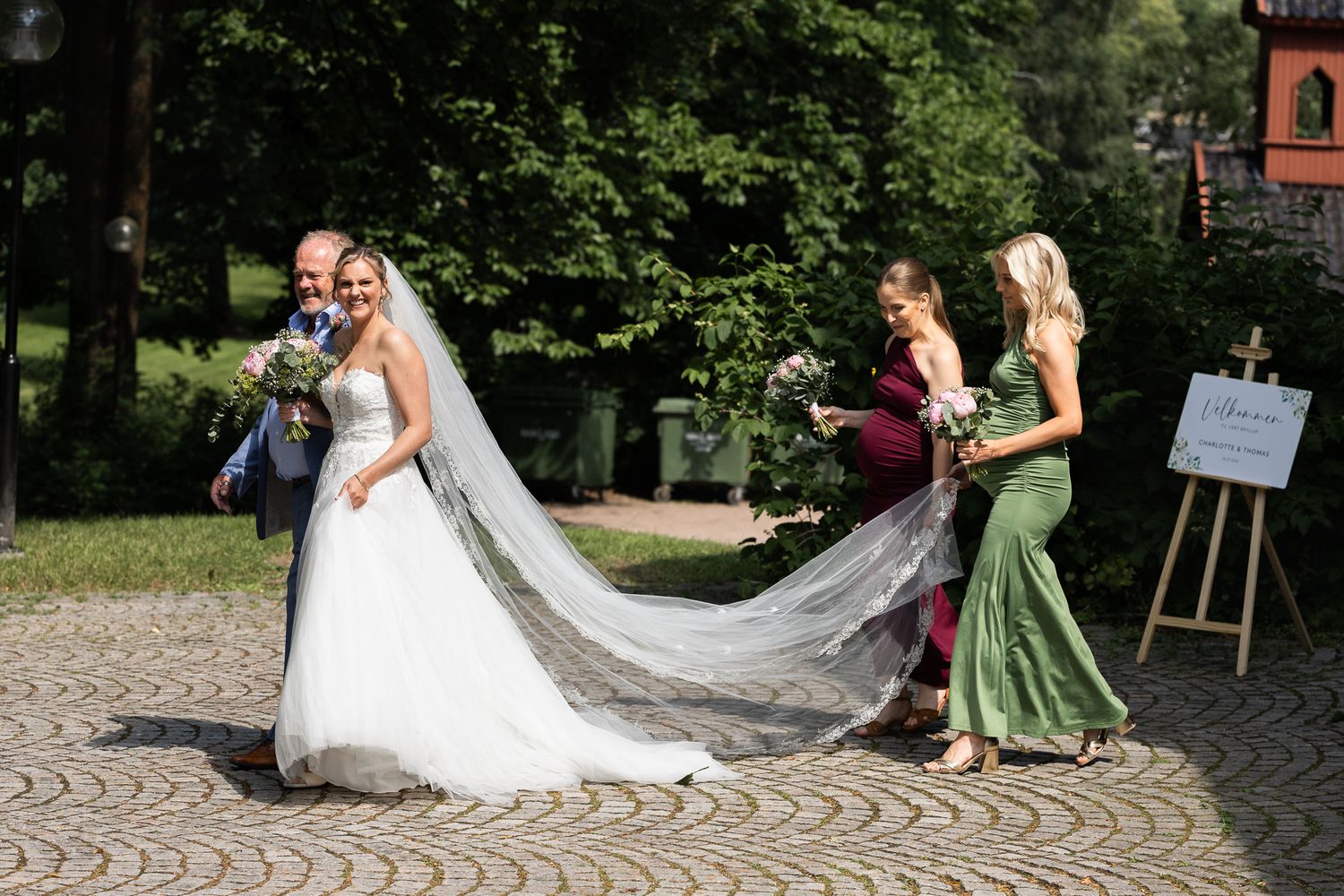 Bride in white gown with long veil held by bridesmaids in green and burgundy dresses outdoors at wedding venue.