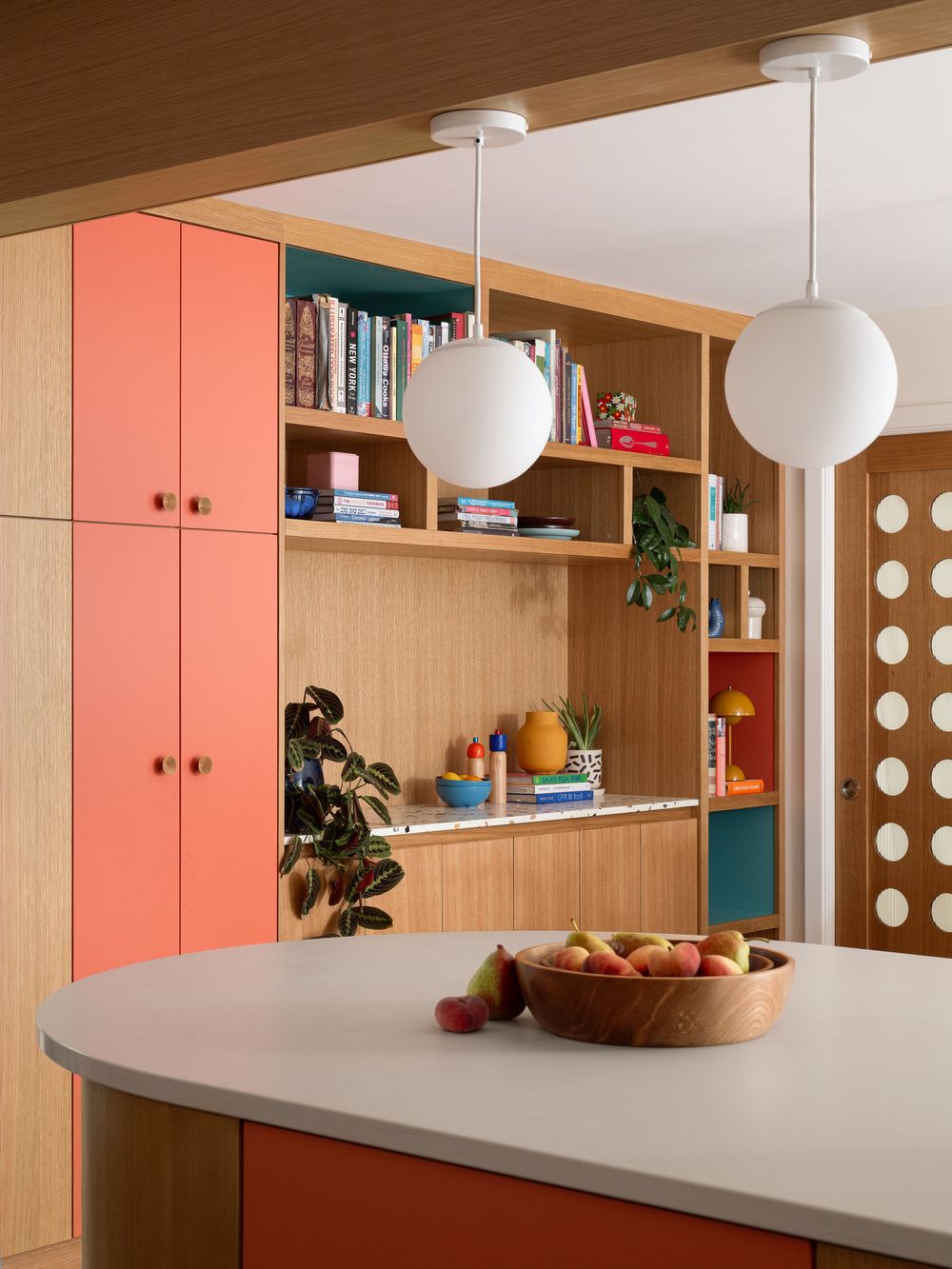 Modern kitchen with coral cabinets, wood shelving, globe pendant lights, and a curved island with fruit bowl.
