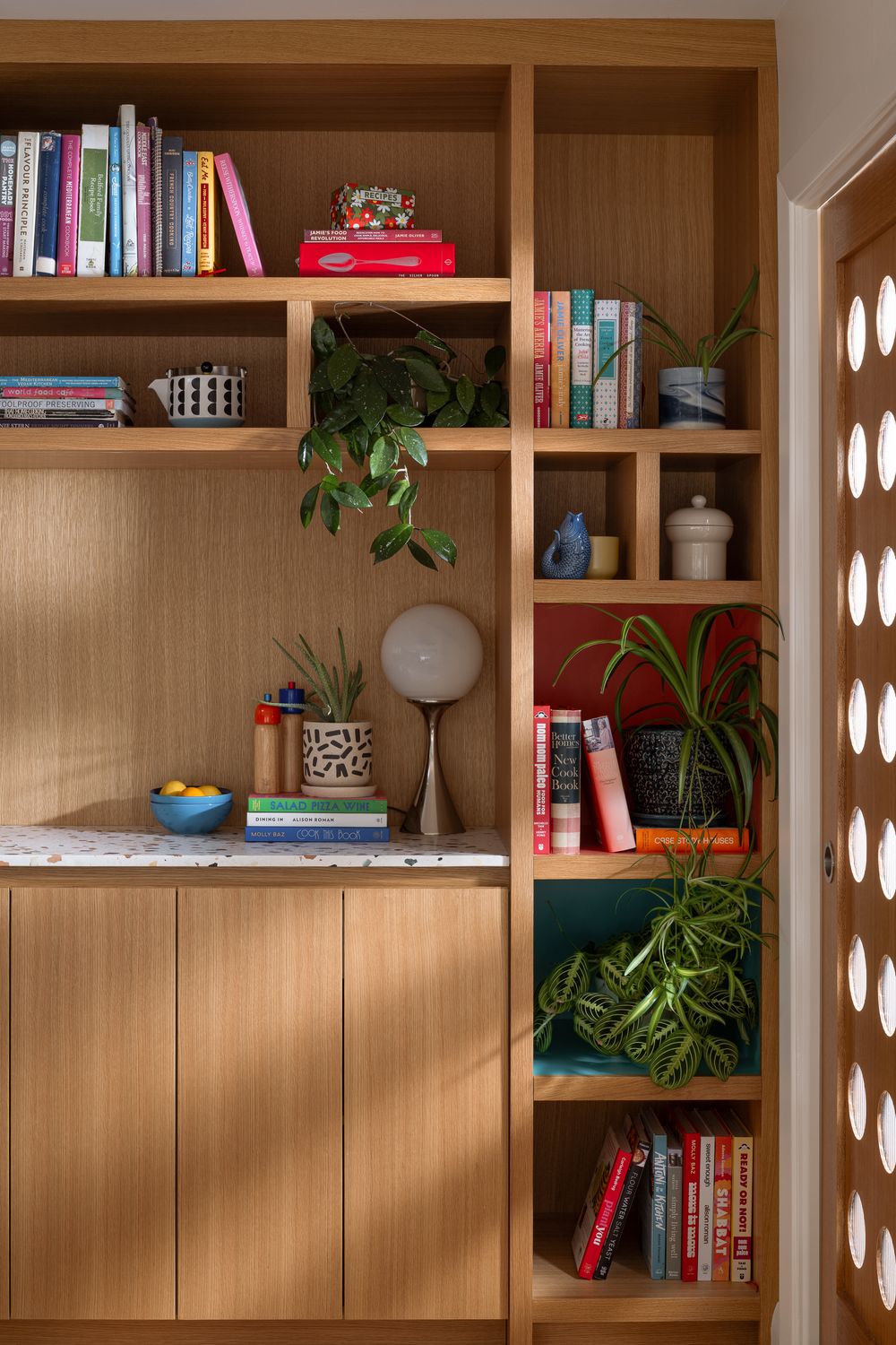 Modern wooden built-in bookshelf styled with colorful books, green plants, and decorative objects in a bright living space.
