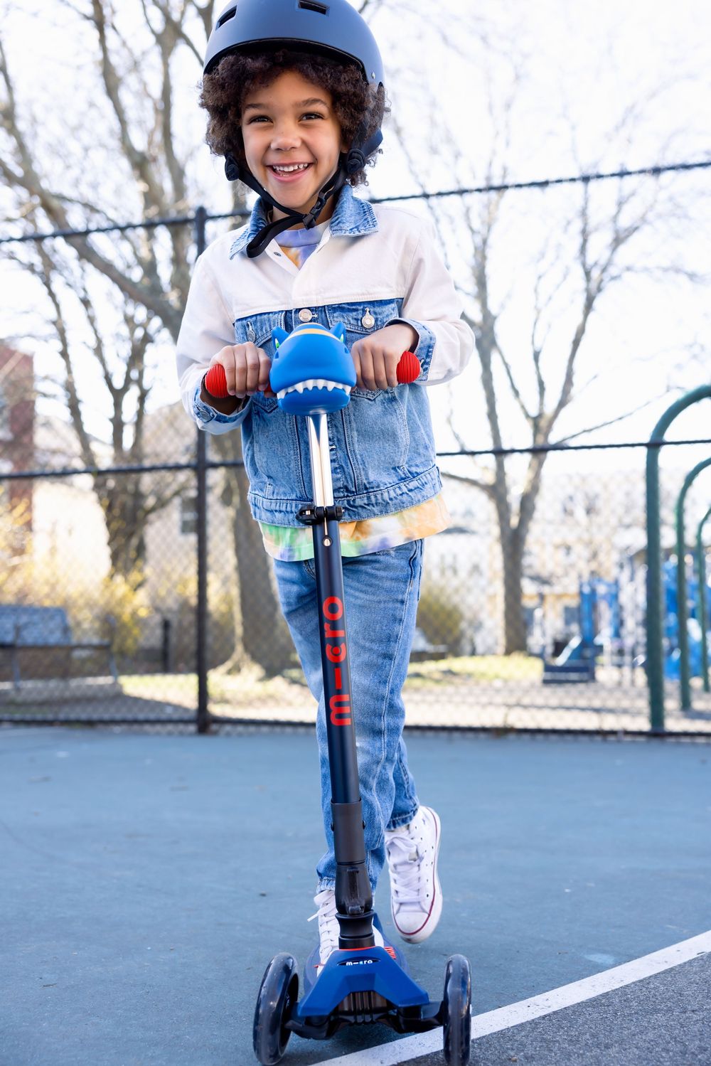 Smiling child wearing a blue helmet rides a blue three-wheel Micro scooter on an outdoor court.