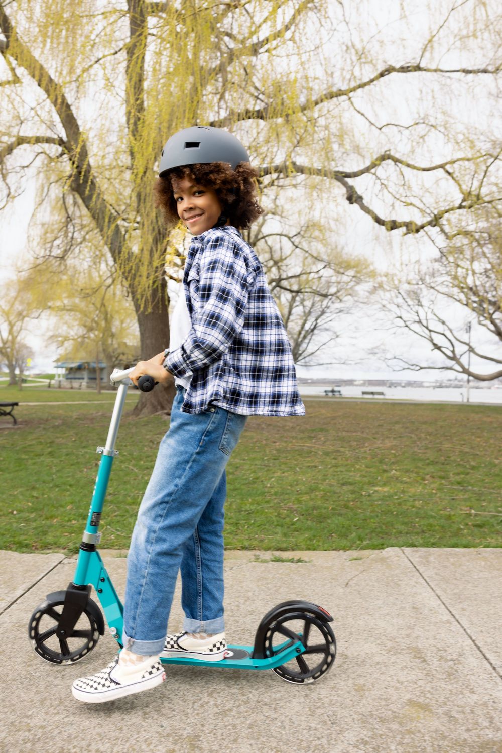 Happy child in a plaid shirt and helmet poses on a teal kick scooter in a park near water.
