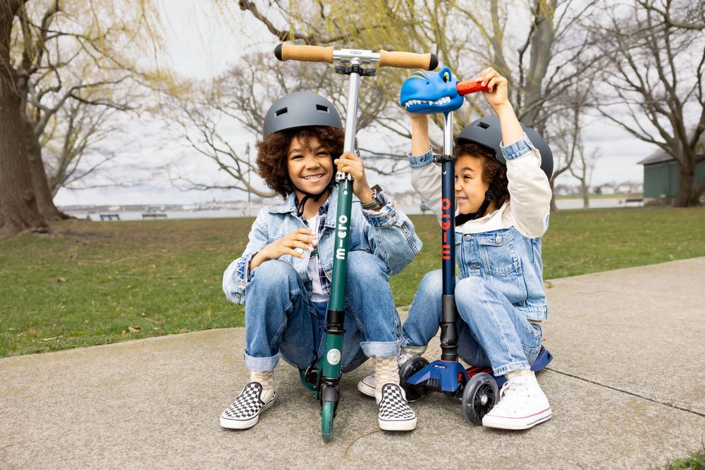 Two smiling kids in helmets crouch beside their Micro scooters in a park setting.