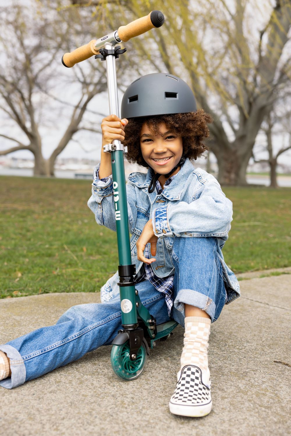 Smiling child in a helmet sits on the ground hugging a green Micro kick scooter outdoors.