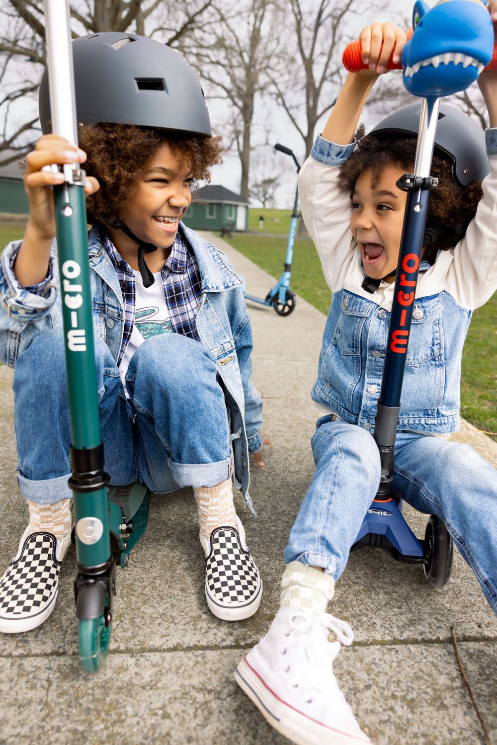 Two kids in denim outfits and helmets laugh while sitting on Ocie scooters on an outdoor pathway.